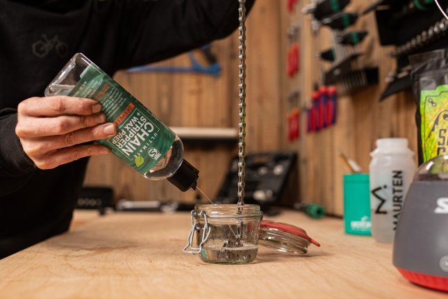 Ein Mechaniker spritzt Antriebsreiniger aus einer durchsichtigen Plastikflasche in ein Weckglas, in welches eine Fahrradkette hängt. 