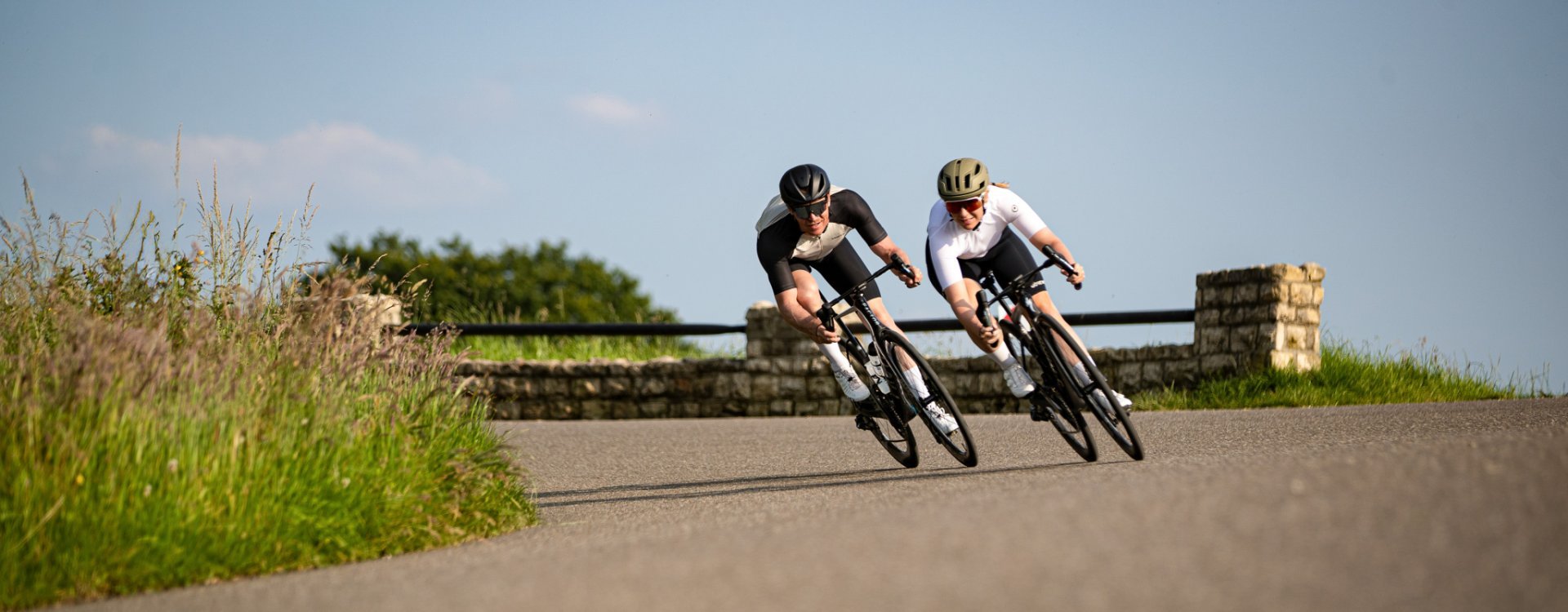 Two road cyclists speed towards the camera while leaning into a turn. Perfect asphalt under a blue sky.