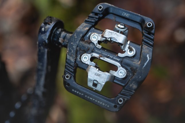 Photo de détail : Un OneUp Clip Pedal noir avec une cage en acier inoxydable. Dans le flou, on distingue un sol forestier automnal.