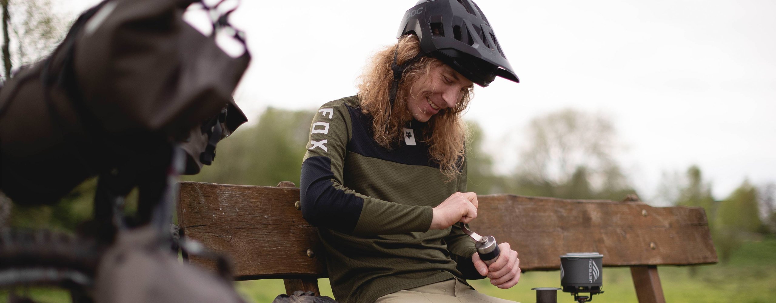 An MTB bikepacker takes a break on a park bench and grinds coffee with a compact coffee grinder