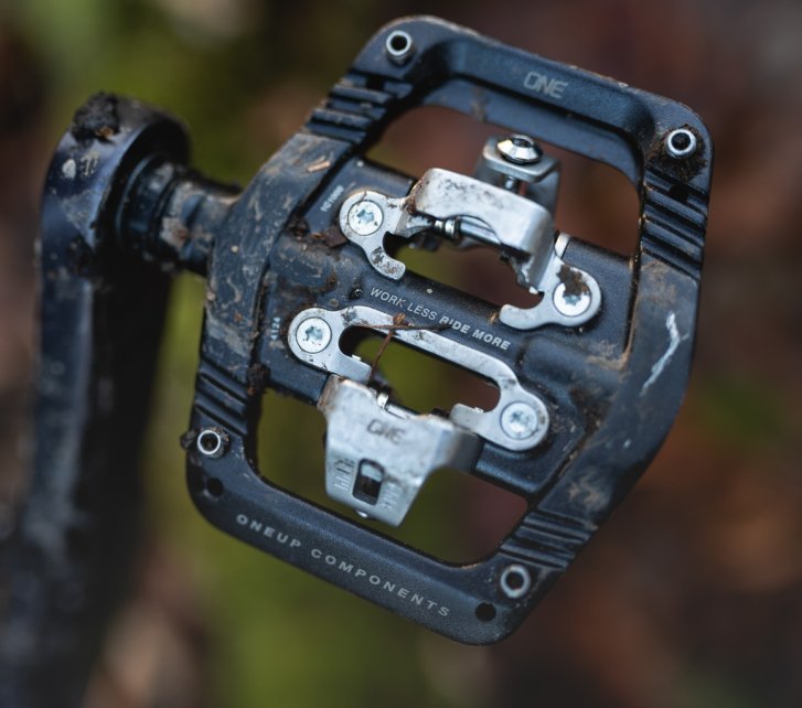 Detailfoto: Ein schwarzes OneUp Clip Pedal mit Edelstahlkäfig. In der Unschärfe ist herbstlicher Waldboden erkennbar.