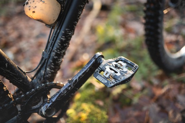 Photo de détail : Un OneUp Clip Pedal noir sur un VTT sombre. On ne voit qu'une manivelle et des parties du tube inférieur. En arrière-plan flou, on distingue le sol forestier automnal et une roue avant partiellement visible.