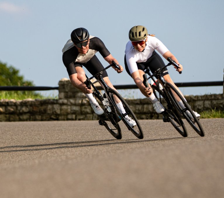 Two road cyclists speed towards the camera while leaning into a turn. Perfect asphalt under a blue sky.