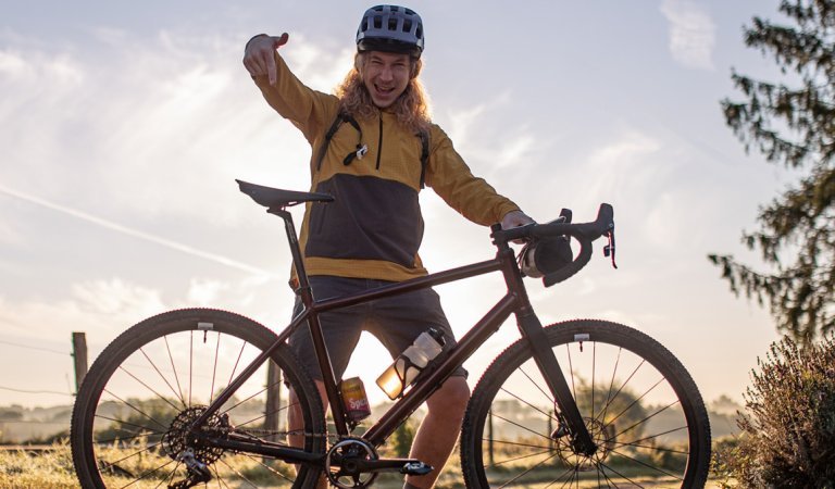 Photo à contre-jour sur un chemin agricole en gravier : Kai, employé de bc, se tient fièrement derrière son nouveau vélo de gravel et le désigne du doigt.