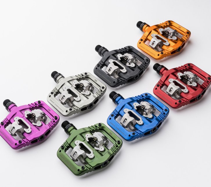 Sieben bunte OneUp Clip Pedals für MTB liegen nebeneinander auf einer weißen Fläche.