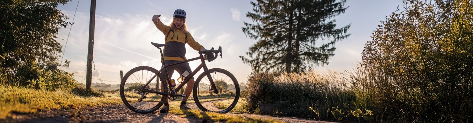 Backlit shot on a gravel farm road: bc employee Kai stands proudly behind his new gravel bike and points at it.