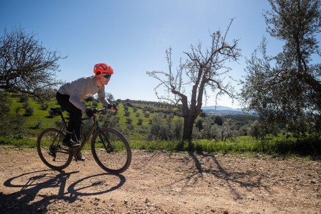 Un homme portant un casque orange roule sur un vélo gravel sur un chemin de gravier, avec un paysage toscan baigné de lumière et un ciel bleu en arrière-plan.