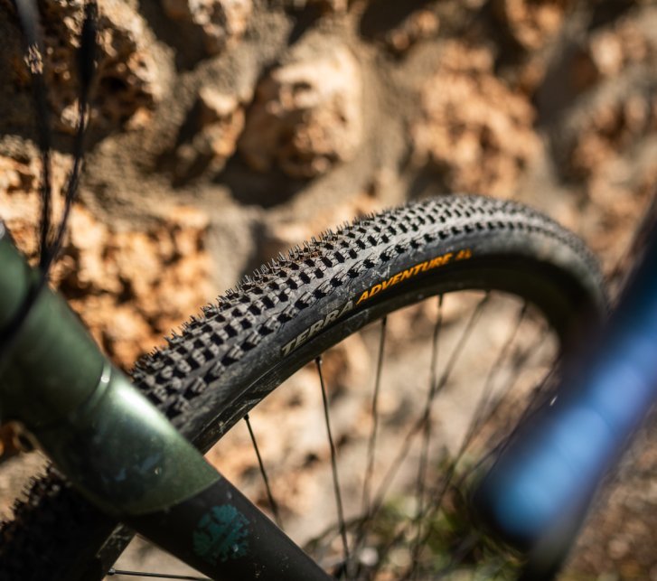 Photo en gros plan d’un pneu gravel à crampons monté à l’avant d’un vélo vert olive, placé devant un mur en pierre naturelle couleur terracotta.