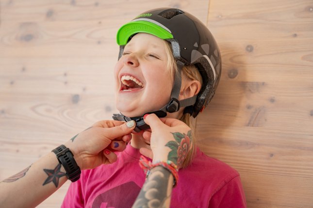 Les mains tatouées et colorées d’une femme adulte ferment la sangle du menton d’un casque de vélo autour du cou d’une fille blonde.