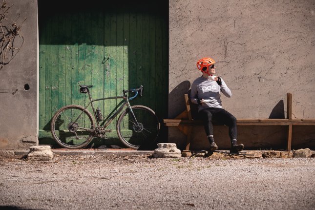 Un vélo gravel est appuyé contre un portail en bois vert. À côté, un homme portant un casque orange est assis sur un banc en bois, une tasse en porcelaine blanche à la main.