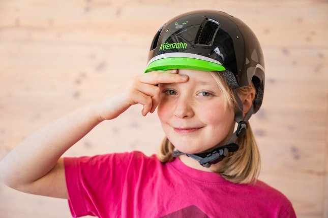 Une fille blonde porte un casque de vélo noir de la marque Affenzahn. Elle place deux doigts sur ses sourcils pour montrer où le casque doit se poser.