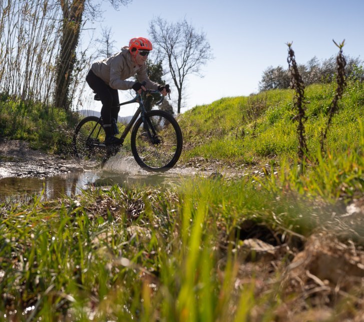 Un homme avec un casque orange traverse un petit ruisseau à vélo gravel, là où le chemin est coupé par l’eau. Les éclaboussures brillent à contre-jour.