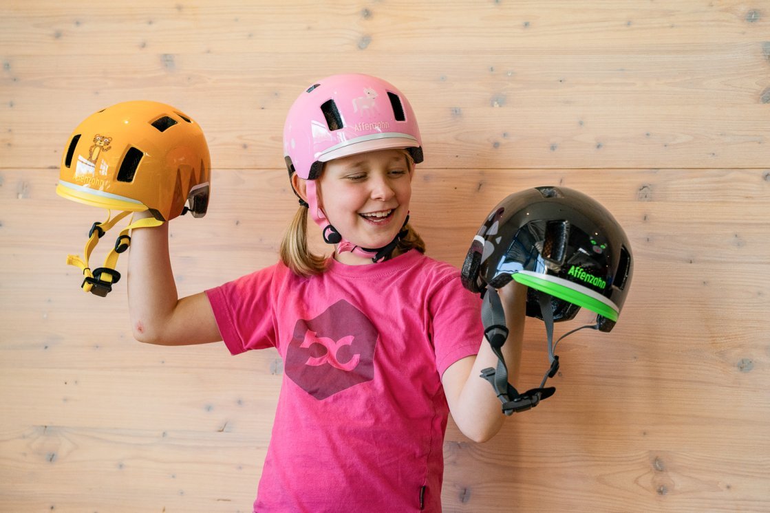 Ein blondes, etwa 10-jähriges Mädchen in einem pinken bc-T-shirtsteht vor einer hellen Holzwand trägt einen rosafarbenen Fahrradhelm und hält in jeder Hand einen weiteren Fahrradhelm.