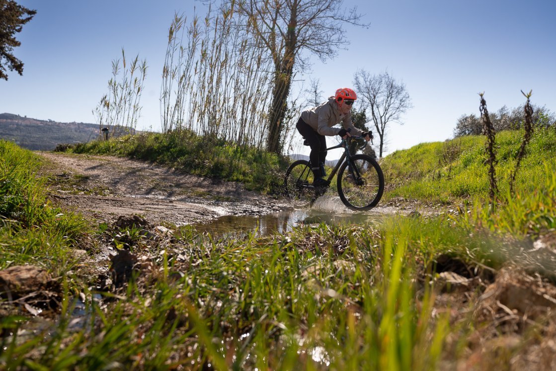 Un homme avec un casque orange traverse un petit ruisseau à vélo gravel, là où le chemin est coupé par l’eau. Les éclaboussures brillent à contre-jour.