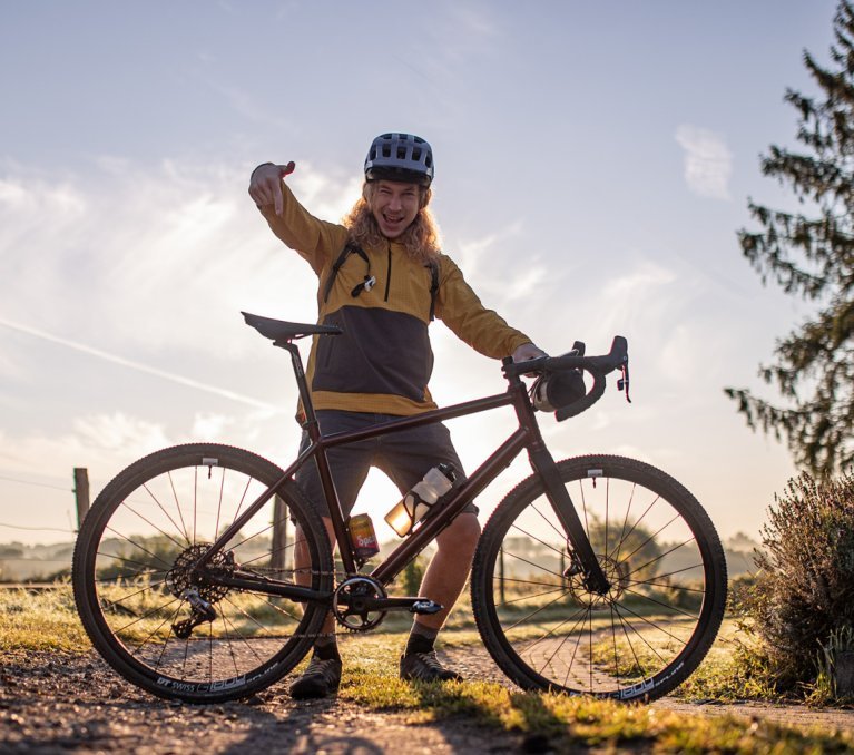 Backlit shot on a gravel farm road: bc employee Kai stands proudly behind his new gravel bike and points at it.