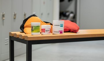 Pictured are 4 varieties of chamois cream from Elite, Assos and Eule in front of 2 bib shorts on a bench in the bc men's locker room.