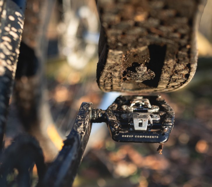 Photo de détail : La semelle d'une chaussure de VTT, positionnée juste au-dessus d'un OneUp Clip Pedal noir. En arrière-plan flou, on distingue des parties d'un VTT sombre ainsi qu'un sol forestier automnal.