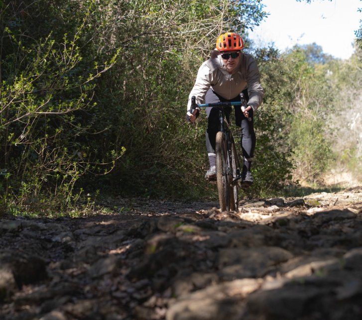 Un homme avec un casque orange roule à vélo gravel vers la caméra sur un chemin accidenté et rocailleux. Des broussailles épaisses bordent le sentier.