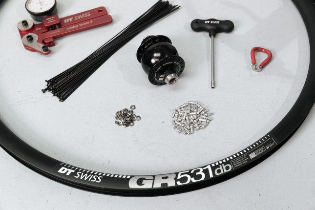 On a white surface, the individual parts and tools for a custom wheel build are laid out: rim, spokes, nipples, hub with dynamo, spoke wrench, and a spoke tension meter.