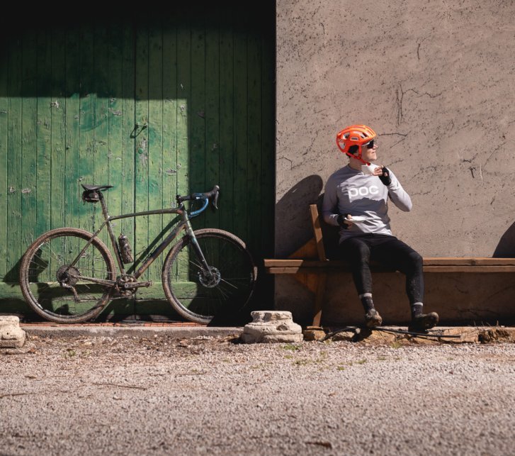 Un vélo gravel est appuyé contre un portail en bois vert. À côté, un homme portant un casque orange est assis sur un banc en bois, une tasse en porcelaine blanche à la main.