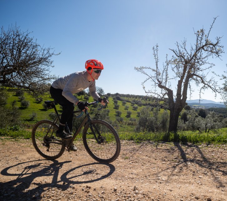 Un homme portant un casque orange roule sur un vélo gravel sur un chemin de gravier, avec un paysage toscan baigné de lumière et un ciel bleu en arrière-plan.