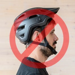 Portrait d'un homme aux cheveux bruns de profil. Il porte un casque de vélo trop en arrière sur le cou, avec un cercle rouge et une barre diagonale superposée à l'image.