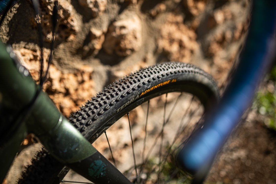 Photo en gros plan d’un pneu gravel à crampons monté à l’avant d’un vélo vert olive, placé devant un mur en pierre naturelle couleur terracotta.