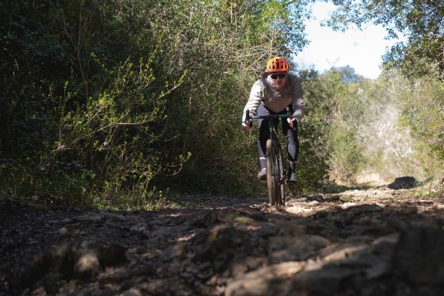 Un homme avec un casque orange roule à vélo gravel vers la caméra sur un chemin accidenté et rocailleux. Des broussailles épaisses bordent le sentier.