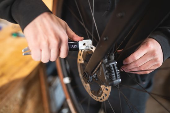 A hand holds an alignment tool against a brake disc mounted on a road bike.