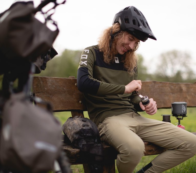 An MTB bikepacker takes a break on a park bench and grinds coffee with a compact coffee grinder