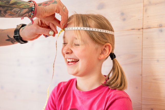 Die tätowierten Hände mit buntem Nagellack einer Mutter halten ein Maßband um den Kopf eines kichernden blonden Kinds.