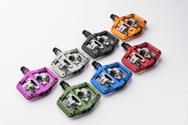 Sept OneUp Clip Pedals colorés pour VTT sont disposés côte à côte sur une surface blanche.