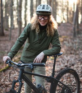 L'employé de bc, Kai, s'appuie décontracté et souriant contre son VTT dans la forêt d'Aix-la-Chapelle. Kai a de longues boucles blondes et porte un casque de VTT clair ainsi qu'une veste de pluie olive.