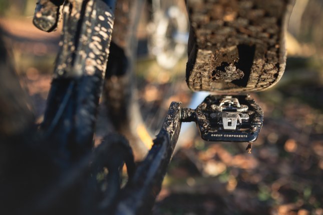 Photo de détail : La semelle d'une chaussure de VTT, positionnée juste au-dessus d'un OneUp Clip Pedal noir. En arrière-plan flou, on distingue des parties d'un VTT sombre ainsi qu'un sol forestier automnal.