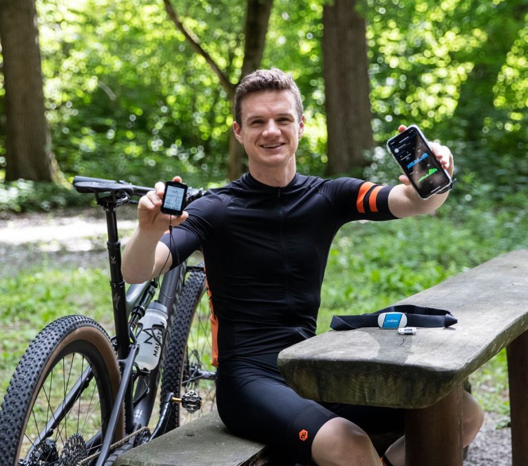 Christof, de l'équipe de gestion des produits de bc, tient son ordinateur de vélo Garmin Edge et son smartphone, qui affiche ses données de performance, face à la caméra.