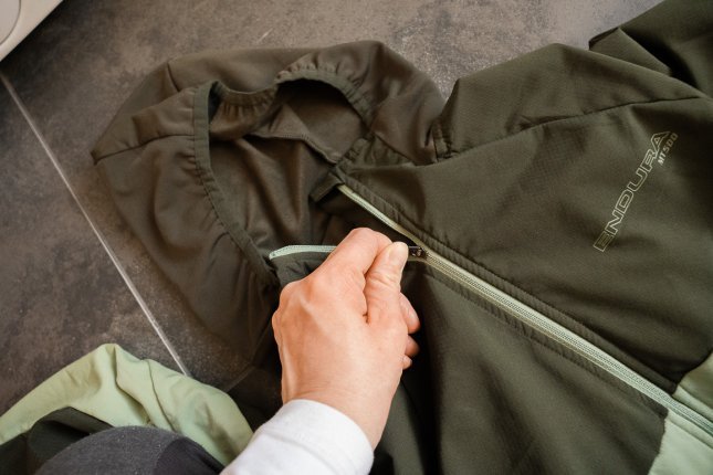 Une veste fonctionnelle verte est posée sur un sol carrelé gris. Une main ferme la fermeture éclair avant.