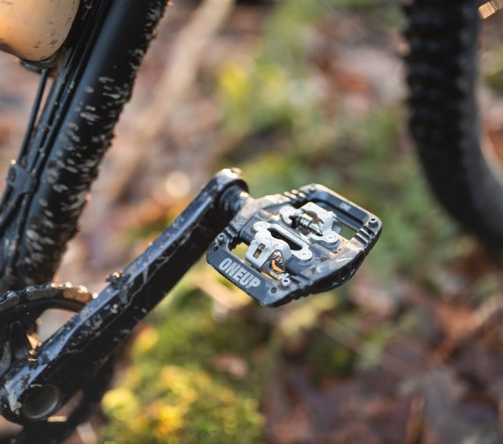 Detailfoto: Ein schwarzes OneUp Clip Pedal an einem dunklen Mountainbike. Wir sehen nur einen Kurbelarm und Teile des Unterrohrs. In der Unschärze herbstlicher Waldboden und ein Vorderrad im Anschnitt.