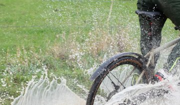 Ein Mann fährt mit einem Fahrrad durch eine tiefe Pfütze. Schlammiges Wasser spritzt zu allen Seiten.